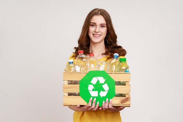 Adolescente positive souriante aux cheveux bruns tenant une boîte de bouteilles en plastique et symbole vert de recyclage tri des déchets économiser l'écologie Prise de vue en studio intérieure isolée sur fond gris