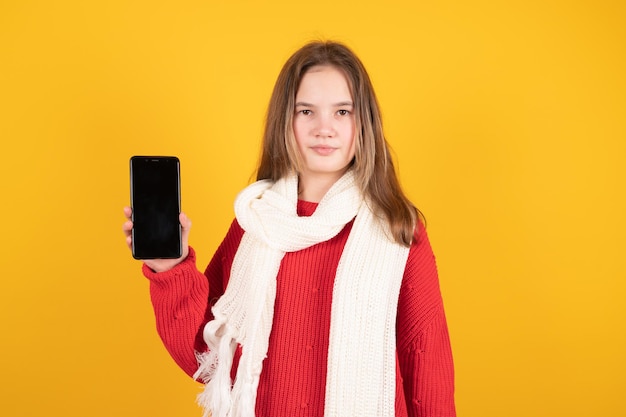 Adolescente en pull chaud et écharpe tenant un téléphone avec écran blanc isolé sur fond de couleur jaune