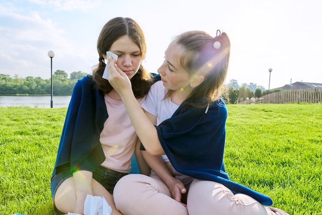 Adolescente qui pleure et petite amie réconfortante, adolescents assis sur l'herbe dans le parc. Amitié, adolescence, santé mentale