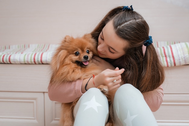 Une adolescente avec une race de chien Spitz se réjouit avec un animal de compagnie à la maison sur le sol.