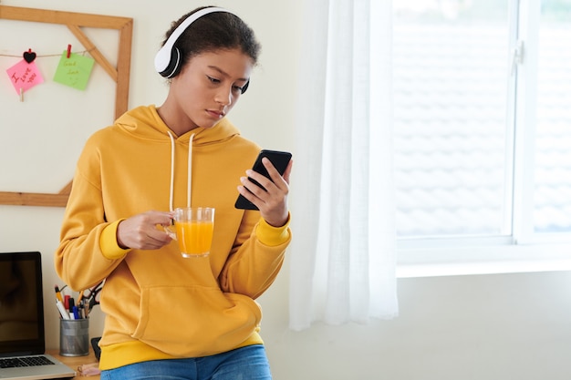 Adolescente de race mixte sérieuse dans des écouteurs sans fil et un sweat à capuche jaune debout dans sa chambre avec des notes adhésives sur le mur et regardant une vidéo sur un smartphone