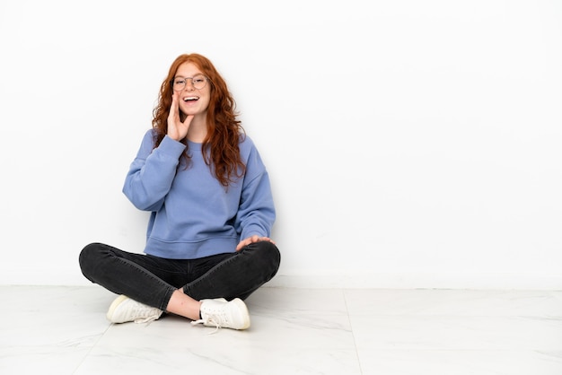 Adolescente rousse assise sur le sol isolée sur fond blanc avec une expression faciale surprise et choquée