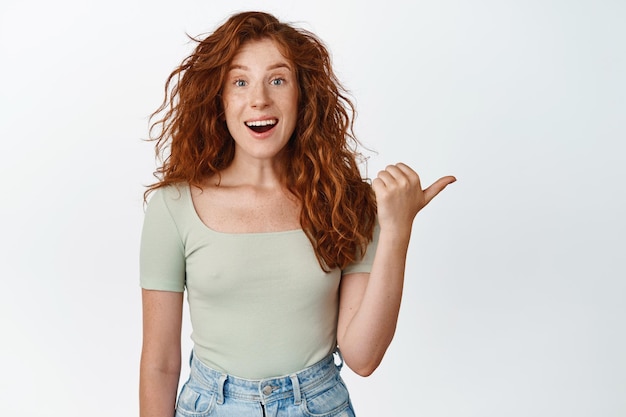 Adolescente rousse excitée pointant le doigt vers la droite en souriant et en regardant étonné de vérifier la vente montrant le logo debout sur fond blanc