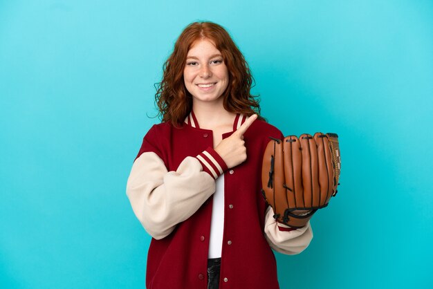 Adolescente rousse avec gant de baseball isolé sur fond bleu pointant vers le côté pour présenter un produit