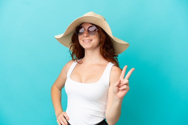 Adolescente rousse isolée sur fond bleu en maillot de bain en vacances d'été et faisant un geste de victoire