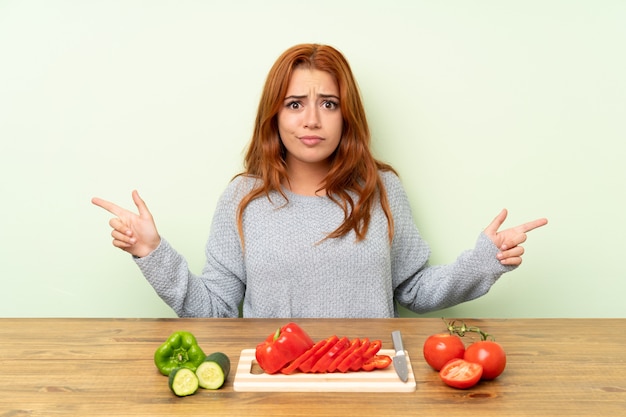 Adolescente rousse avec des légumes dans un tableau pointant vers les latérales ayant des doutes