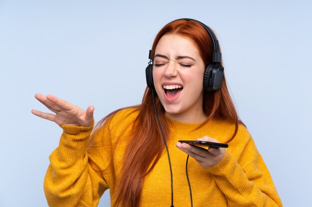 Adolescente rousse sur mur bleu isolé écouter de la musique avec un mobile et chanter