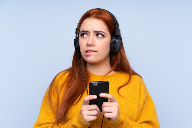 Adolescente rousse sur mur bleu isolé écouter de la musique avec un mobile et penser