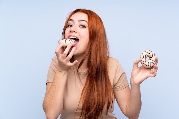 Adolescente rousse sur mur bleu isolé manger un beignet