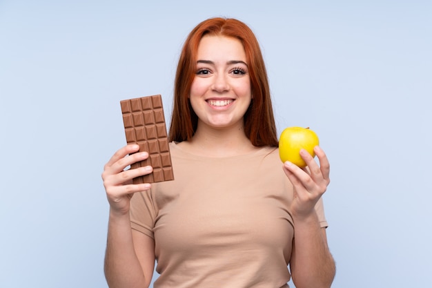 Adolescente rousse sur mur bleu isolé en prenant une tablette de chocolat dans une main et une pomme dans l'autre