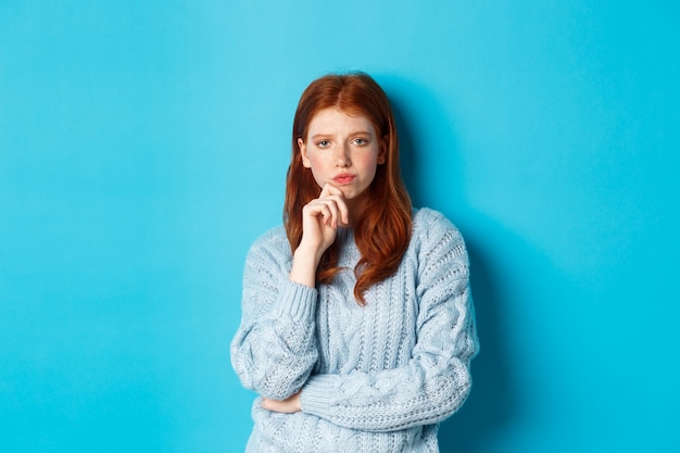 Une adolescente rousse réfléchie regardant la caméra, réfléchissant à des idées, prenant une décision avec un visage sérieux, debout sur fond bleu.