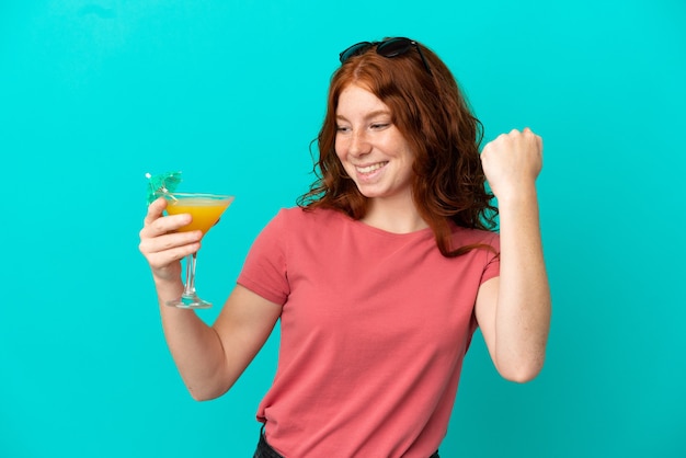 Adolescente rousse tenant un cocktail isolé sur fond bleu célébrant une victoire