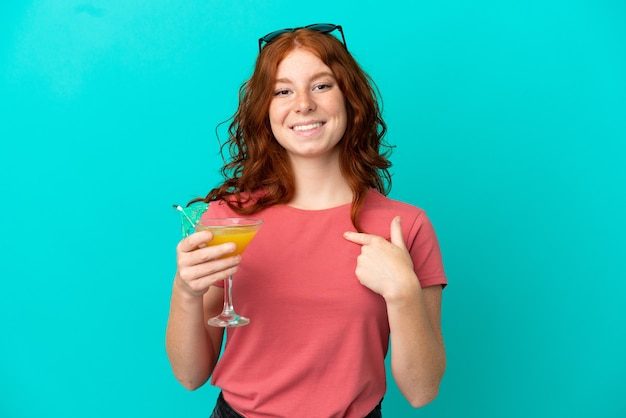 Adolescente rousse tenant un cocktail isolé sur fond bleu avec une expression faciale surprise