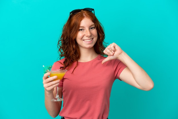 Adolescente rousse tenant un cocktail isolé sur fond bleu fier et satisfait de lui-même