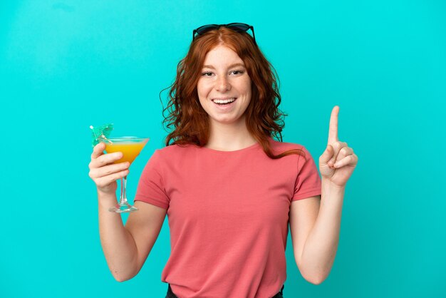 Adolescente rousse tenant un cocktail isolé sur fond bleu pointant vers une excellente idée