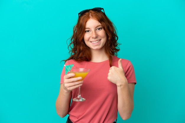 Adolescente rousse tenant un cocktail isolé sur fond bleu avec les pouces vers le haut parce que quelque chose de bien s'est produit