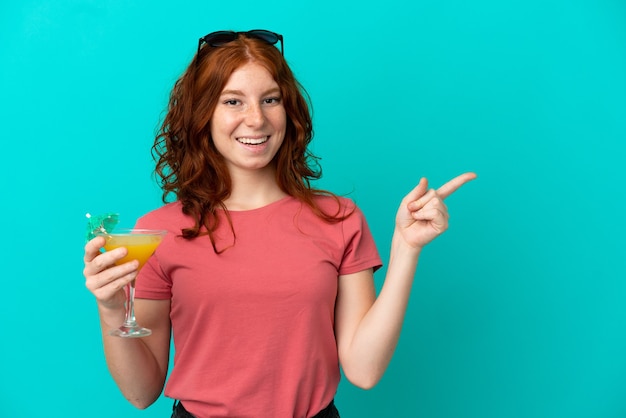 Adolescente rousse tenant un cocktail isolé sur fond bleu surpris et pointant le doigt sur le côté