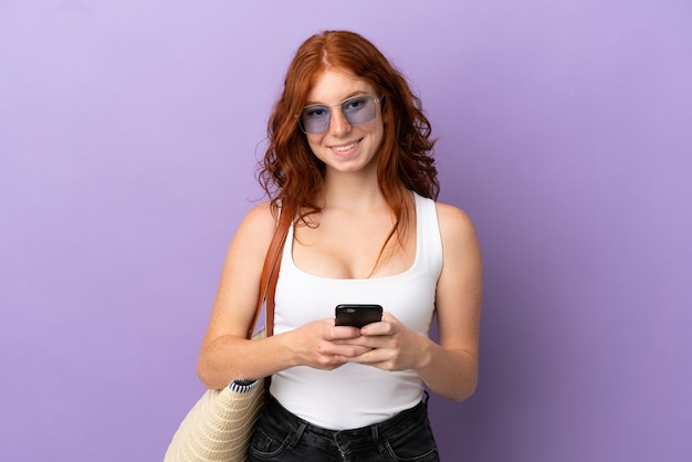 Adolescente rousse tenant un sac de plage isolé sur fond violet envoyant un message avec le mobile