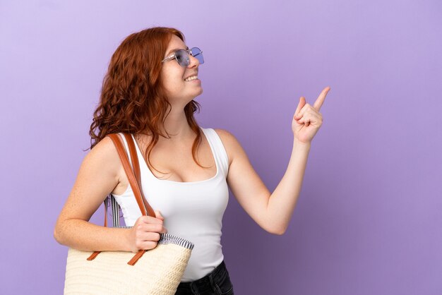 Adolescente rousse tenant un sac de plage isolé sur fond violet, pointant le doigt sur le côté et présentant un produit