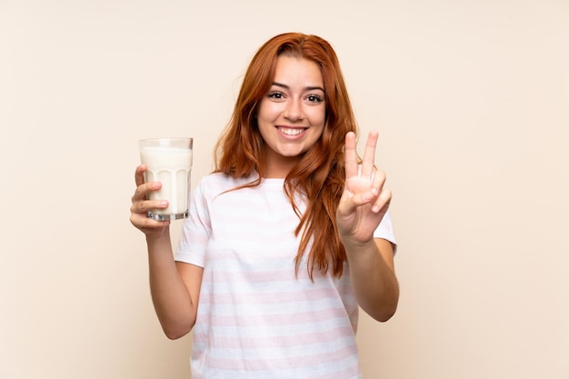 Adolescente rousse tenant un verre de lait sur isolé souriant et montrant le signe de la victoire