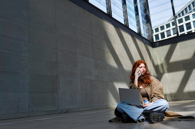 Adolescente rousse utilisant un ordinateur portable parlant sur une cellule dans un emplacement urbain