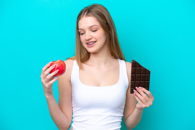Adolescente russe isolée sur fond bleu prenant une tablette de chocolat dans une main et une pomme dans l'autre