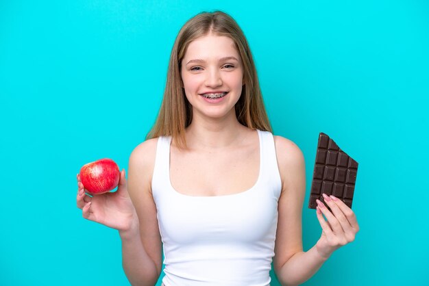 Adolescente russe isolée sur fond bleu prenant une tablette de chocolat dans une main et une pomme dans l'autre