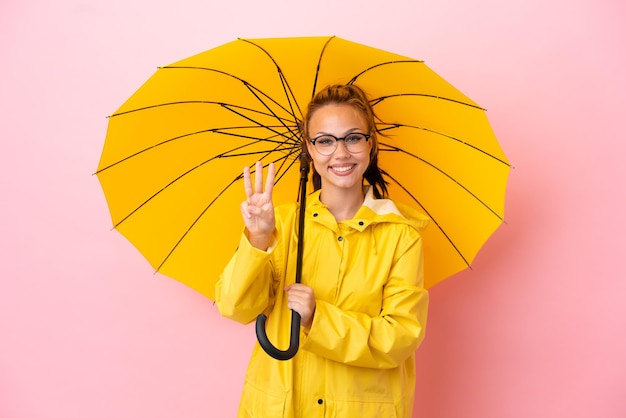 Adolescente russe avec manteau imperméable et parapluie isolé sur fond rose heureux et comptant trois avec les doigts