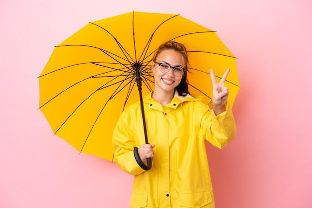 Adolescente russe avec manteau imperméable et parapluie isolé sur fond rose souriant et montrant le signe de la victoire