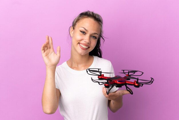 Adolescente russe tenant un drone isolé sur fond violet saluant avec la main avec une expression heureuse