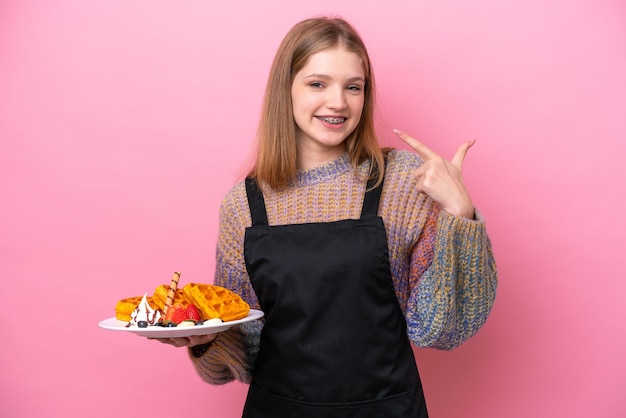 Adolescente russe tenant une gaufre isolée sur fond rose donnant un geste du pouce levé