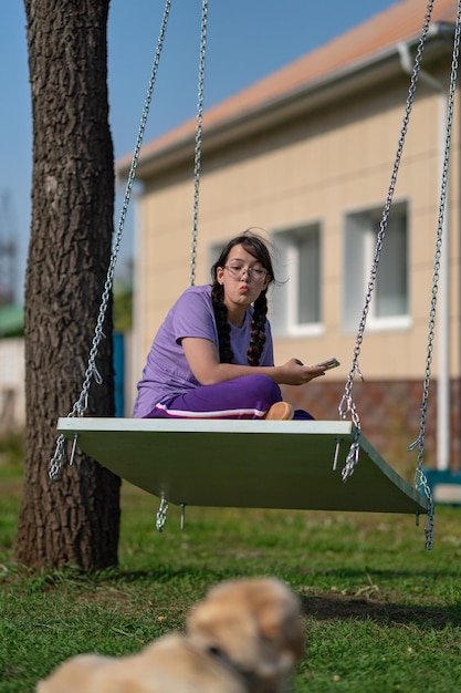 une adolescente se balance sur une balançoire avec un téléphone dans les mains, communique sur les réseaux sociaux