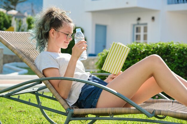 Une adolescente se reposant allongée sur une chaise extérieure sur la pelouse, un livre de lecture féminin, boit de l'eau de la bouteille. Détente, mode de vie, vacances, concept jeunesse
