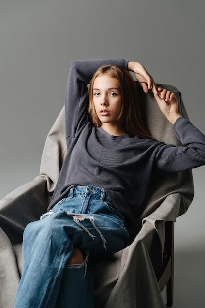 Adolescente sensuelle assise sur un fauteuil, tenant les mains au-dessus de sa tête. Portrait. Porter un jean mom avec des trous et des manches longues grises.