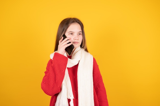 Adolescente sérieuse en écharpe tricotée blanche chaude parlant sur un téléphone portable isolé sur fond jaune