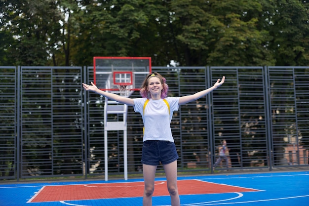 Adolescente souriante active, à la mode avec des cheveux violets, sur un terrain de basket de rue avec les bras levés. Émotion de bonheur, de vacances, de plaisir