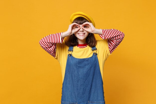 Adolescente souriante en béret français et robe d'été en denim tenant la main près des yeux, imitant des lunettes ou des jumelles isolées sur fond jaune. Émotions des gens, concept de style de vie. Maquette de l'espace de copie.