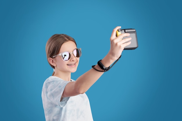 Adolescente souriante à lunettes de soleil et t-shirt tenant une photocamera devant elle-même tout en faisant selfie sur fond bleu