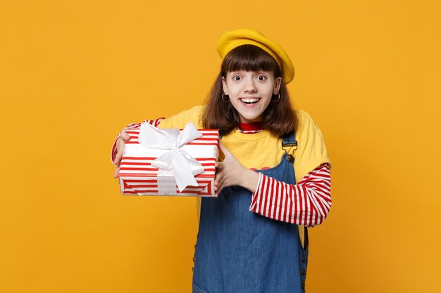 Adolescente surprise en béret français, robe d'été en jean tenant une boîte cadeau rayée rouge avec ruban cadeau isolé sur fond de mur jaune. Concept de vacances d'anniversaire de style de vie d'émotions sincères de personnes.