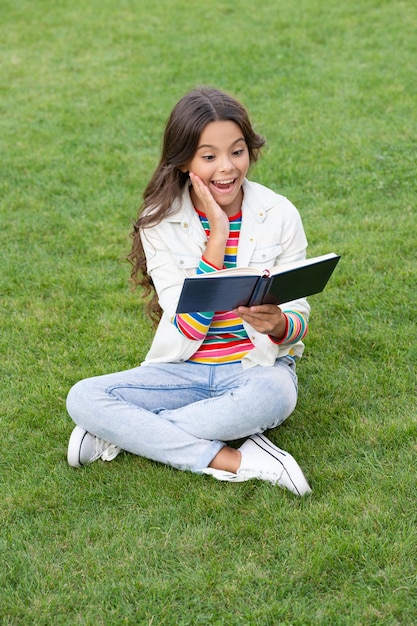 Une adolescente surprise lit un livre assis sur l'herbe en lisant une lectrice de livre fille avec un livre en plein air