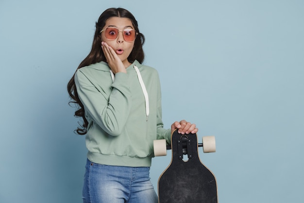 Adolescente surprise à lunettes de soleil tenant une planche à roulettes. La fille de planche à roulettes touche ses joues sur un fond bleu