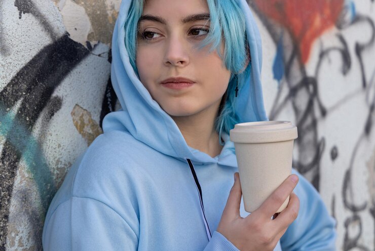 Photo adolescente en sweat à capuche surdimensionné bleu clair avec café à emporter et regardant de côté adolescente aux cheveux bleus restant à l'extérieur près du mur de graffitis maquette de coupe concept hipster et adolescence
