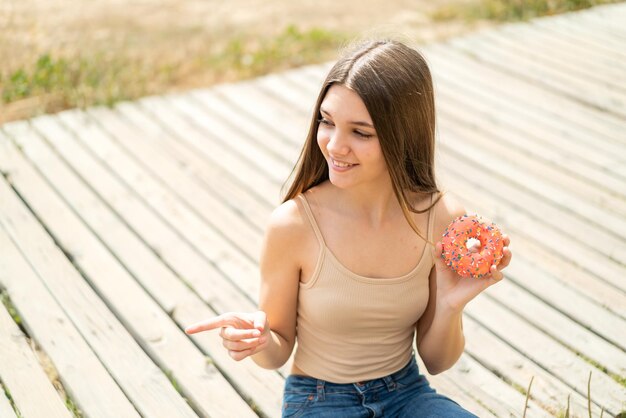 Adolescente tenant un beignet à l'extérieur pointant vers le côté pour présenter un produit
