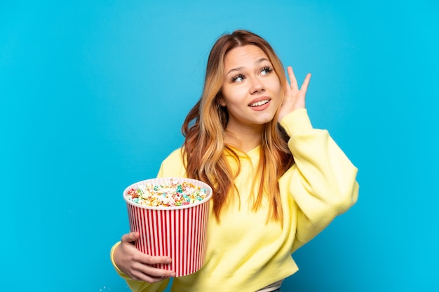 Adolescente tenant des pop-corns sur fond bleu isolé écoutant quelque chose en mettant la main sur l'oreille