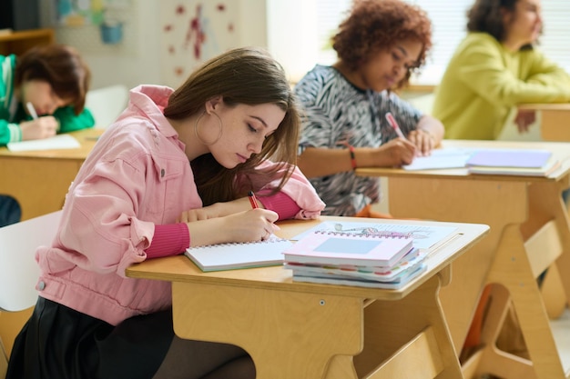 Adolescente en tenue décontractée prenant des notes dans un cahier tout en étant assise près du bureau