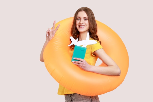 Adolescente touristique optimiste en T-shirt jaune debout avec un anneau en caoutchouc orange, tenant un document de passeport et une maquette d'avion, se réjouissant de la tournée de voyage. Tourné en studio intérieur isolé sur fond gris.
