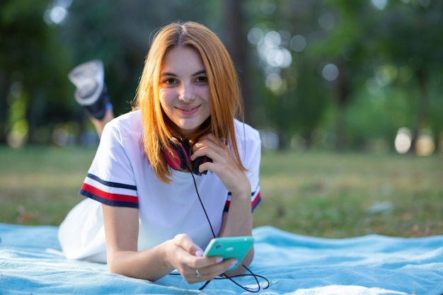 Adolescente très souriante aux cheveux rouges à l'aide de sellphone à l'extérieur dans le parc.