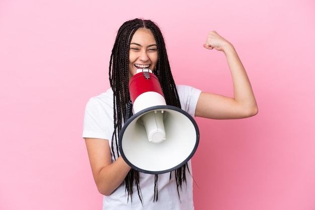 Adolescente avec des tresses sur fond rose isolé criant à travers un mégaphone pour annoncer quelque chose