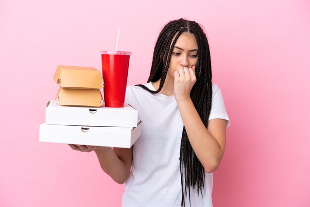 Adolescente avec des tresses tenant des pizzas et des hamburgers sur fond rose isolé ayant des doutes