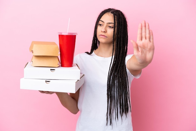 Adolescente avec des tresses tenant des pizzas et des hamburgers sur fond rose isolé faisant un geste d'arrêt et déçu